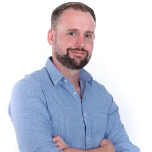 A man with beard and blue shirt standing in front of white background.